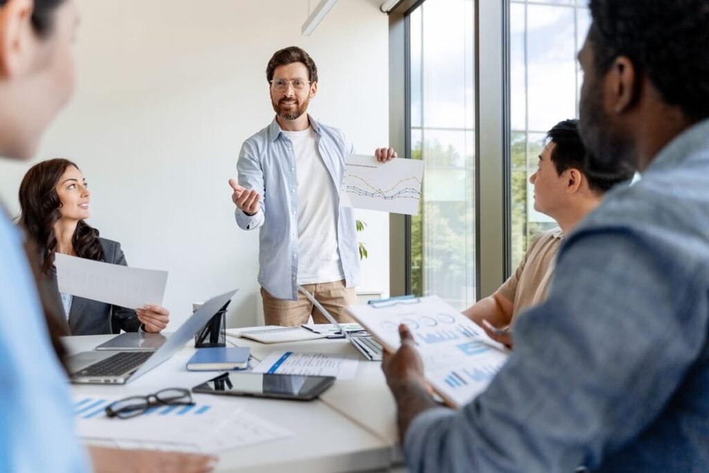Modernūs pardavimai biure – verslo susitikimas, kuriame vyras pristato augimo grafiką komandai, akcentuodamas šiuolaikinį požiūrį į klientą.